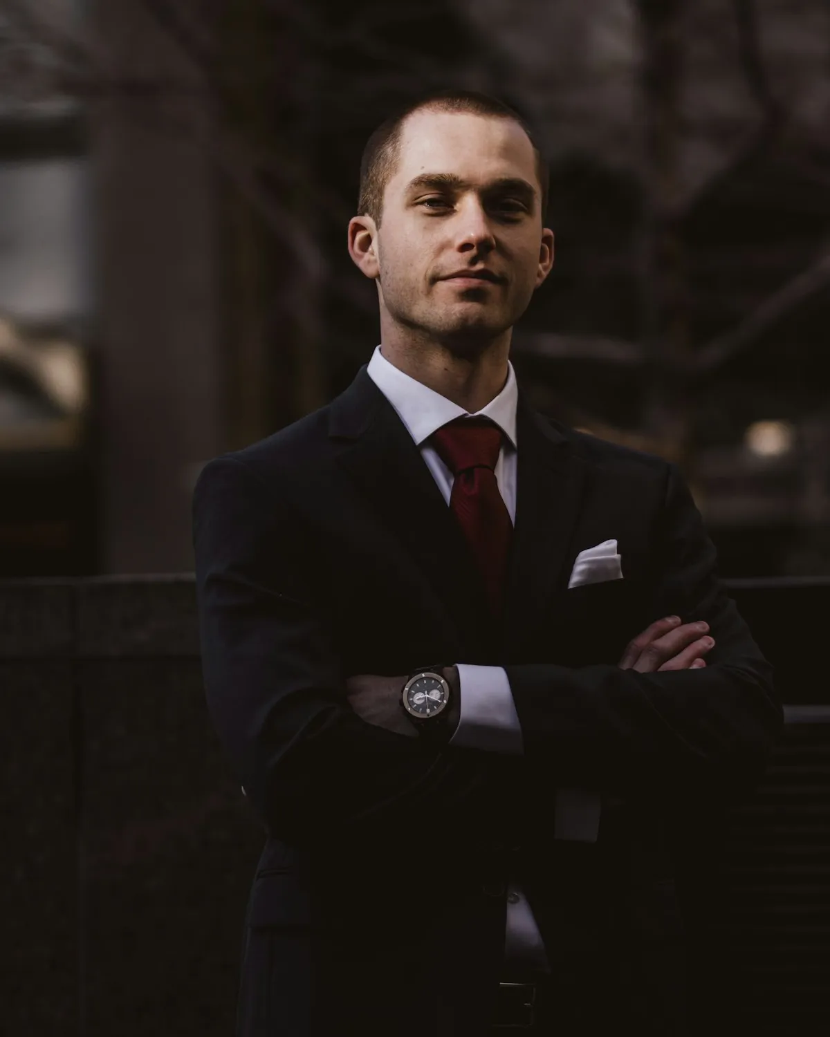 A high-contrast black and white editorial portrait of a photographer in a tailored dark coat, arms crossed, dramatic shadows against a blurred urban backdrop.