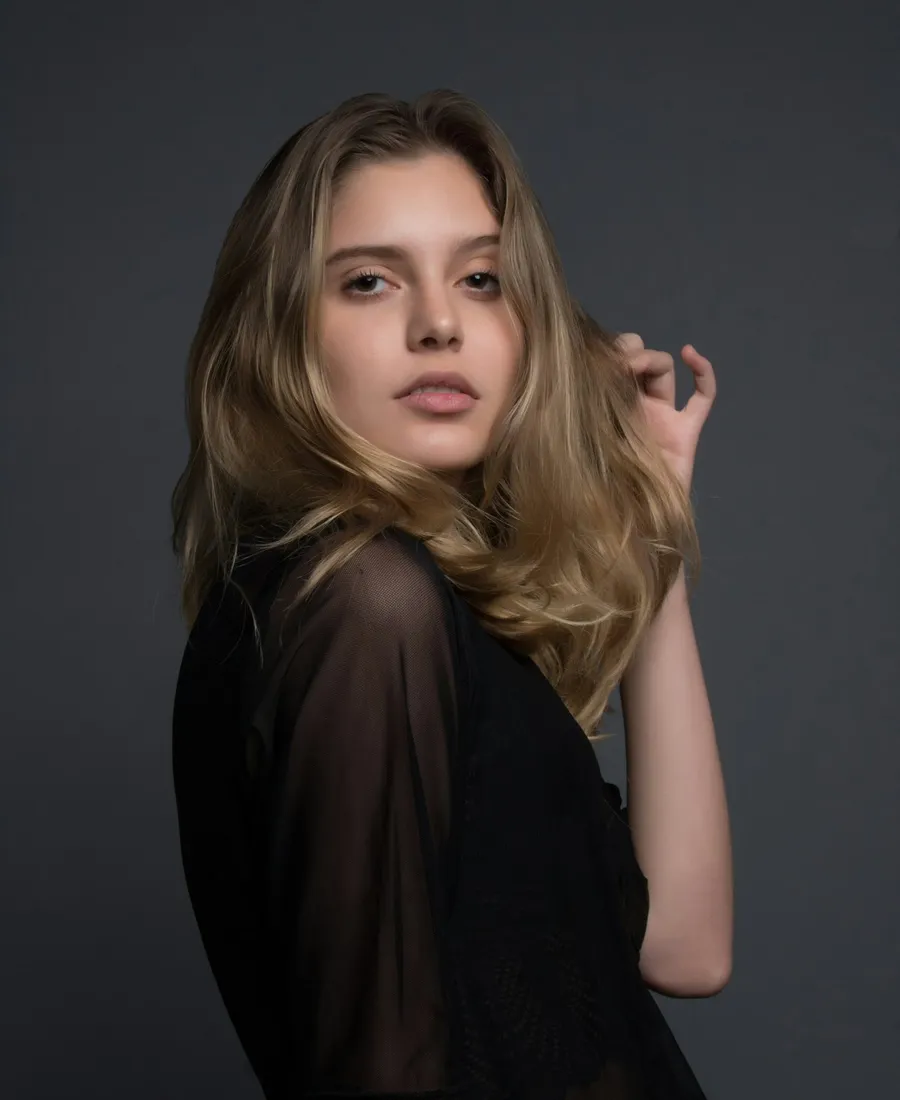 A moody black and white close-up portrait of a woman in a sheer dark top against a plain backdrop, soft dramatic lighting reminiscent of film noir.